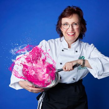 brand portrait of dietician holding a bowl and whisking pink liquid and standing against a blue background
